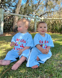 Two children wearing matching shirts and pants set , Terry cloth fabric , sitting outdoors on a grassy area with trees in the background.