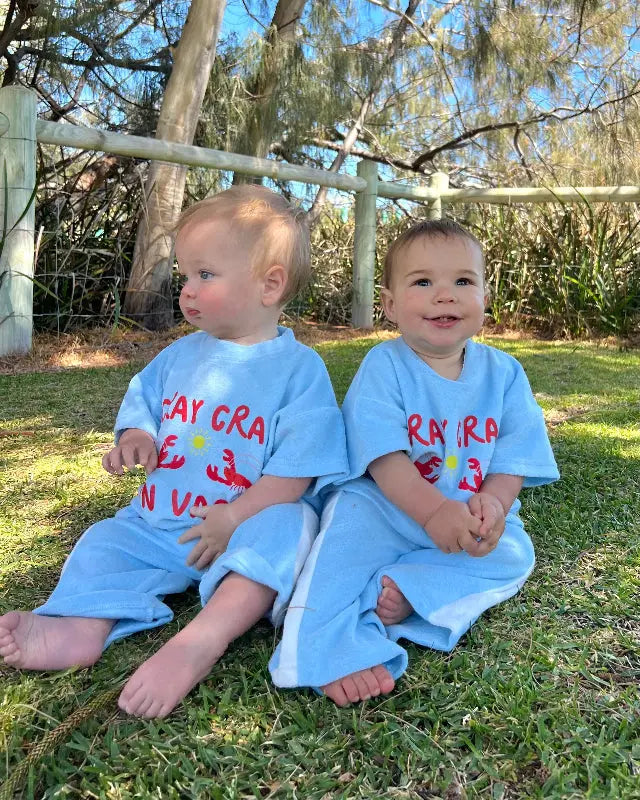 Two children wearing matching shirts and pants set , Terry cloth fabric , sitting outdoors on a grassy area with trees in the background.