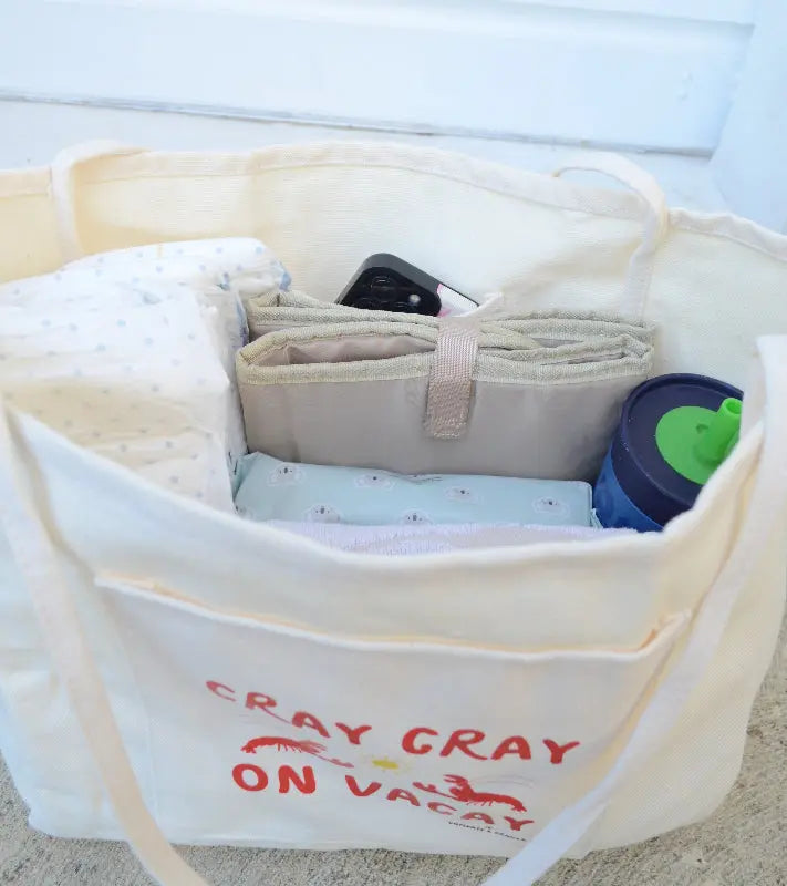 Canvas Beige tote bag with 'Cray Cray on Vacay' text, containing folded clothes and a blue container.