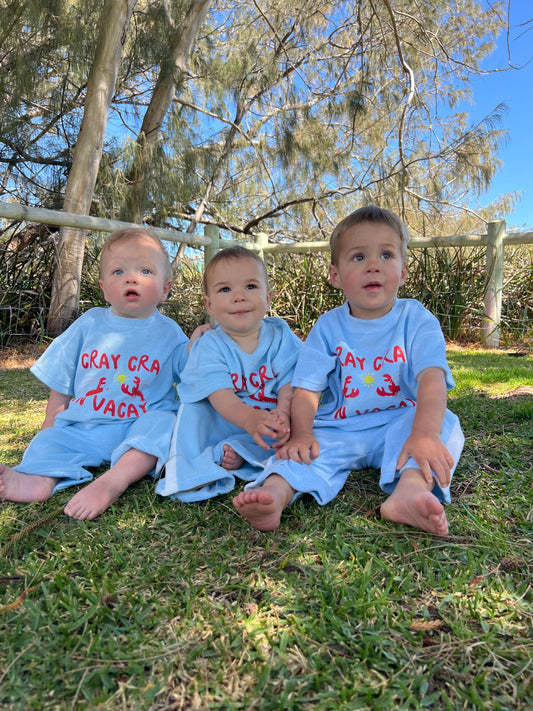 Three children wearing matching shirts and pants set , Terry cloth fabric , sitting outdoors on a grassy area with trees in the background.